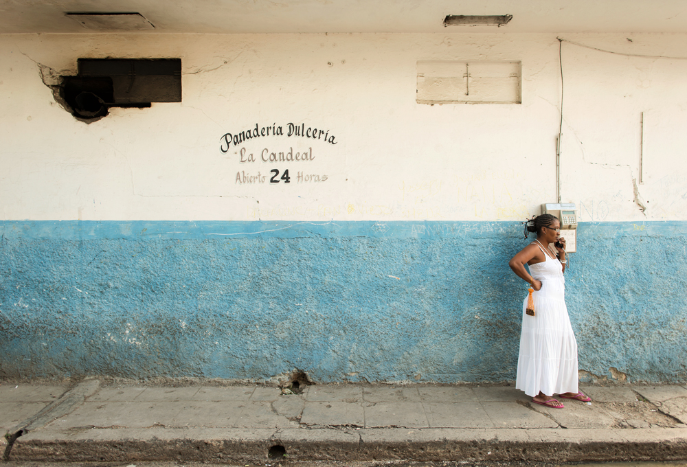 Eine Frau in einem weißen Kleid telefoniert an einem öffentlichen Telefon vor der Fassade der Panadería La Candeal in Havanna, Kuba.