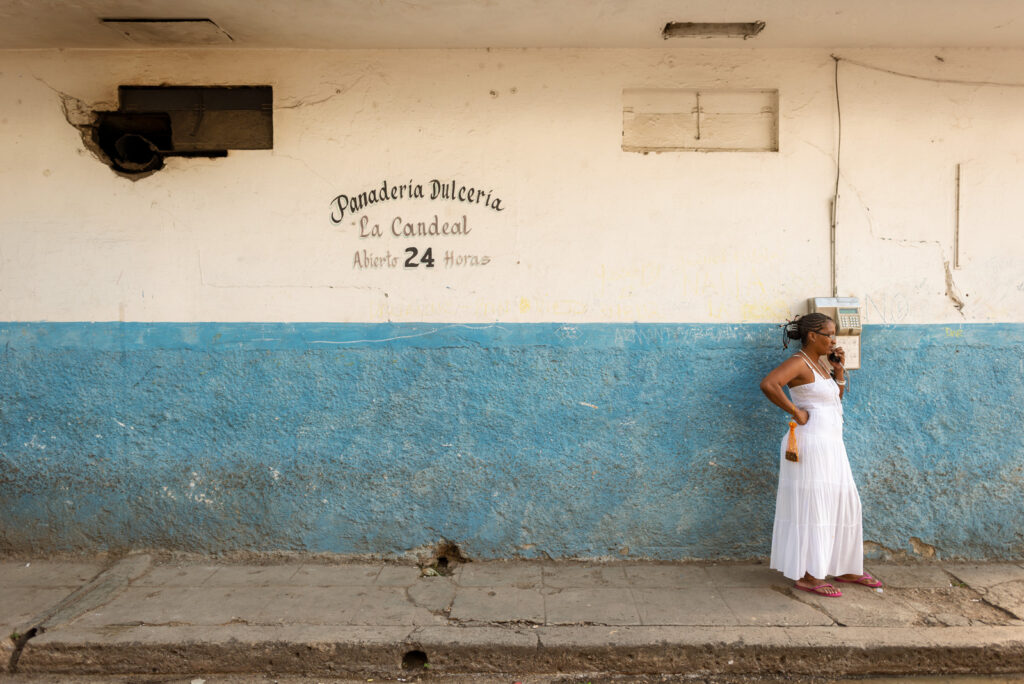 Eine Frau in einem weißen Kleid telefoniert an einem öffentlichen Telefon vor der Fassade der Panadería La Candeal in Havanna, Kuba