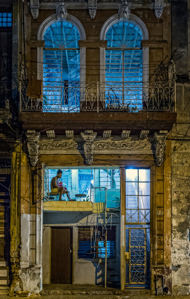 Nächtliche Hausfassade in Havanna mit blau beleuchtetem Innenraum, Balkon und einer Person auf einer offenen Galerie an der Calle Neptuno