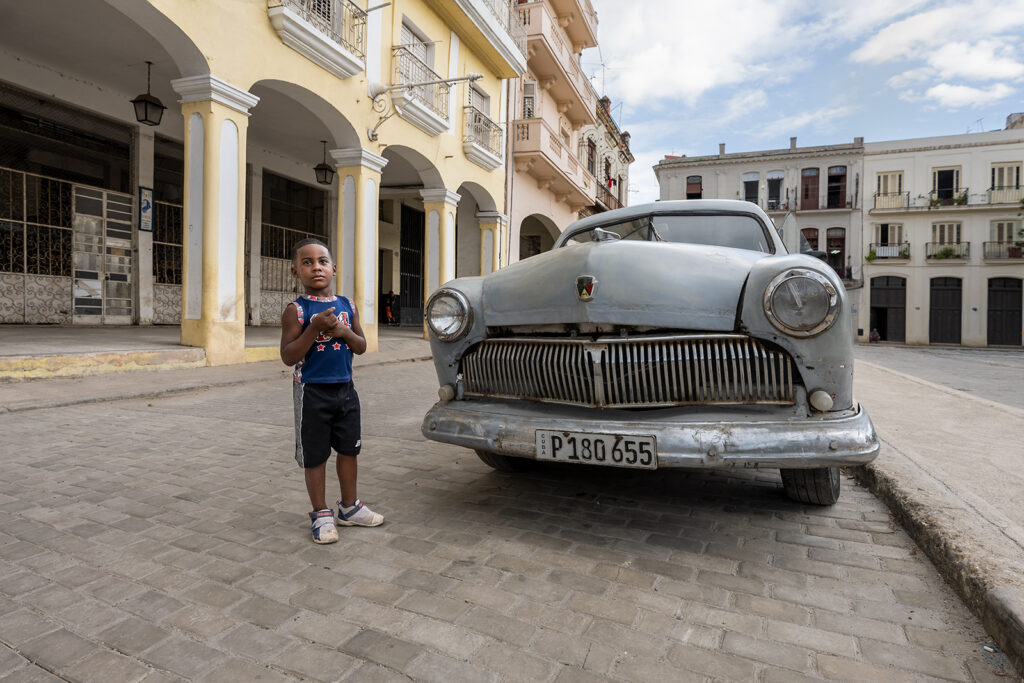 Junge steht neben einem klassischen amerikanischen Chevrolet auf einem Platz in Havanna. Im Hintergrund Arkaden und historische Gebäude im Morgenlicht