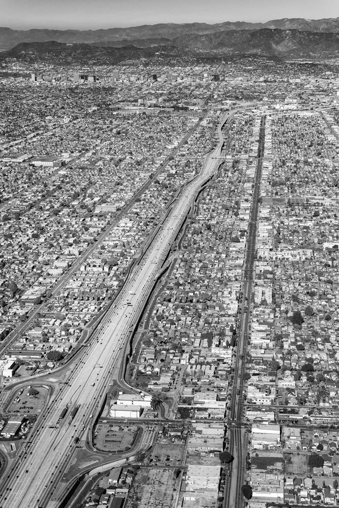 Schwarz-Weiß-Luftaufnahme von Los Angeles mit einer mehrspurigen Autobahn, dicht bebauten Wohngebieten und den San Gabriel Mountains im Hintergrund.