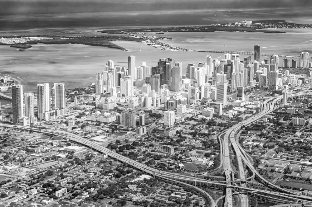 Schwarz-Weiß-Luftaufnahme der Skyline von Miami mit der Biscayne Bay, Brücken und mehrspurigen Verkehrsachsen aus der Perspektive eines startenden Flugzeugs.
