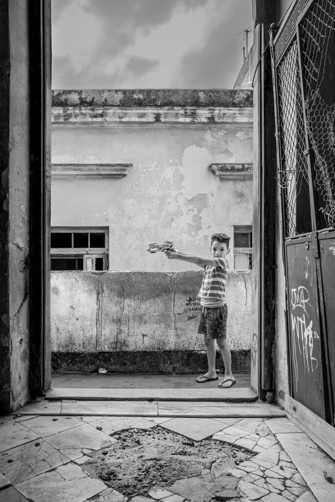 Junge mit Spielzeugpistole in einer Türöffnung eines verfallenen Gebäudes in Havanna, Kuba. Im Vordergrund ein beschädigter Fliesenboden, im Hintergrund verwitterte Fassaden