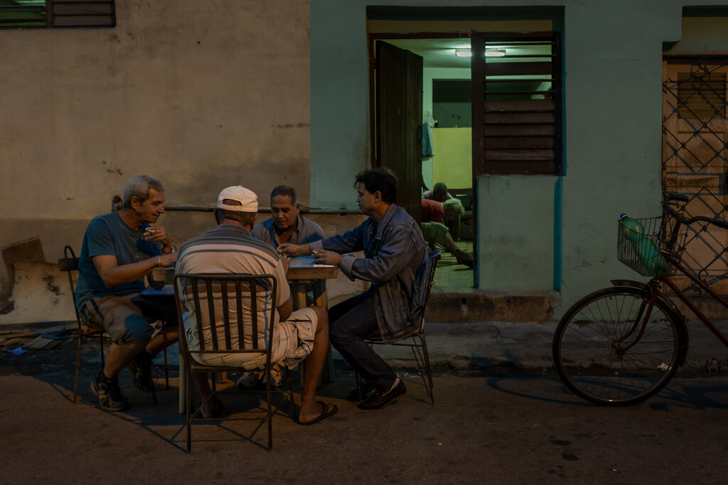 Vier Männer spielen am Abend Domino vor einem Wohnhaus in Havanna. Das warme Licht der Straße und die kühle Beleuchtung aus dem Inneren des Hauses schaffen eine ruhige, alltägliche Atmosphäre