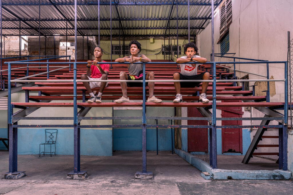 Drei Jugendliche sitzen auf den Rängen einer Boxarena in Havanna und blicken ruhig in die Kamera. Die geometrischen Linien der Tribüne und das weiche Tageslicht verleihen der Szene eine stille, zeitlose Atmosphäre